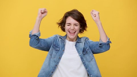 Smiling overjoyed beautiful brunette young woman in denim jacket white t-shirt posing doing winner gesture, say Yes isolated on yellow background in studio. People sincere emotions, lifestyle concept
