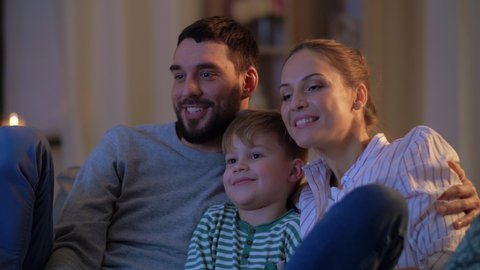 family, leisure and people concept - happy smiling father, mother with remote control and little son watching tv at home at night