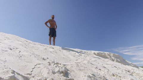 Sexy black milliennial man looks from ontop of sand dune onto beach under blue sky in Australia. Medium to long shot on 4K RED camera.