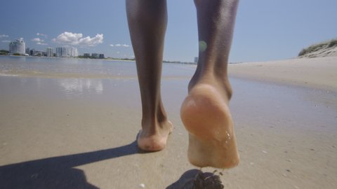 Black male legs and feet walk on sandy beach under blue sky in Australia. Medium to long shot on 4K RED camera.