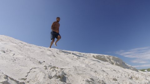 Sexy black milliennial man runs on sand dune on beach under blue sky in Australia. Medium to long shot on 4K RED camera.