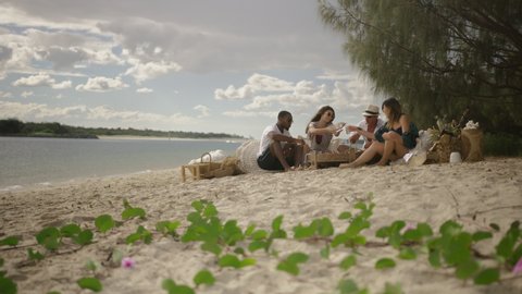 Hungry millennial friends run and to have picnic on sandy beach under sunny blue sky in Australia. Long wide shot on 4K RED camera.