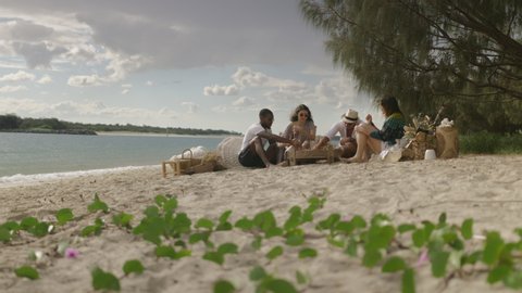 Fun millennial friends run and sit on sandy beach chairs under sunny blue sky in Australia. Long wide shot on 4K RED camera.