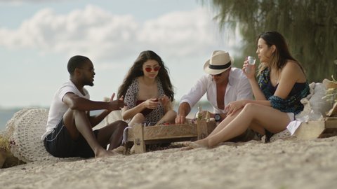 Hungry millennial friends have picnic on sandy beach under sunny blue sky in Australia. Long wide shot on 4K RED camera.