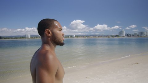 Sexy shirtless black man walks on beach under sunny blue sky in Australia. Medium to closeup on 4K RED camera with lens flare.