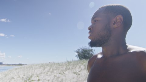 Fit black man looks onto beach under sunny blue sky in Australia. Medium to closeup on 4K RED camera.