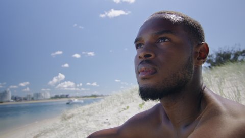 Sexy shirtless black man sits on beach under sunny blue sky in Australia. Medium to closeup on 4K RED camera with lens flare.
