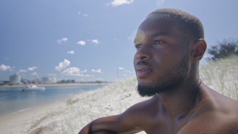 Fit shirtless black man sits on beach under sunny blue sky in Australia. Medium to closeup on 4K RED camera with lens flare.