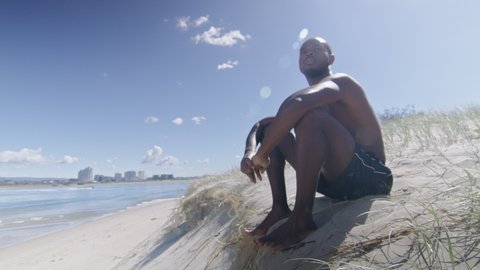 Fit shirtless black man sits on beach under sunny blue sky in Australia. Medium to closeup on 4K RED camera.