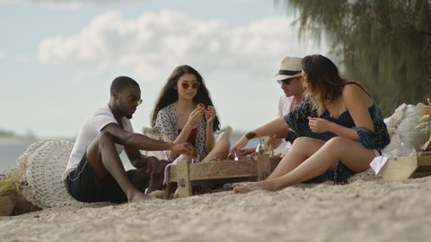 Four millennial friends have picnic on sandy beach under sunny blue sky in Australia. Long wide shot on 4K RED camera.