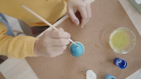 Top view shot of little girl applying dots with paint brush on blue dyed egg while decorating food for Easter