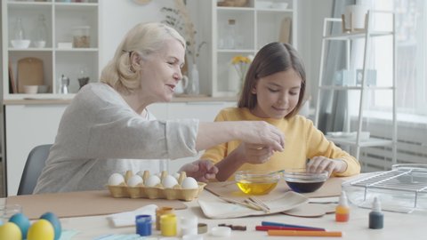 Cute girl dipping egg into bowl with blue food coloring and dying it with help of joyous grandmother while preparing for Easter at home