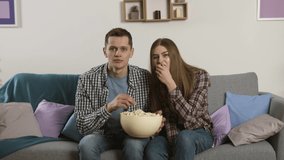 Focused couple watching tv and throwing popcorn to camera with upset face expression