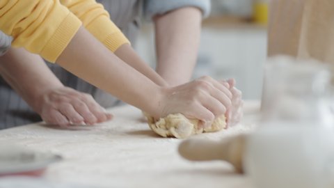 Tilt up shot of little girl in apron smiling and kneading dough on kitchen table with assistance of helpful grandmother