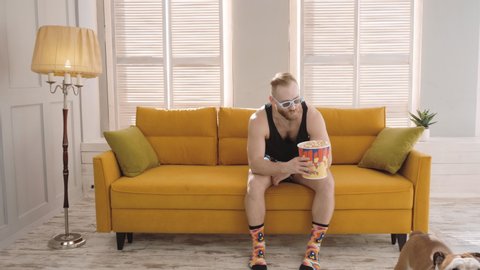 Man and a bulldog at home, sitting on a bright yellow sofa, watching a movie with 3 d glasses, eating popcorn. Love for the pet. The concept of lifestyle and pets.