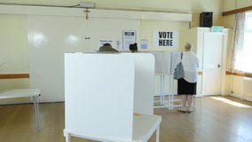 Senior Caucasian Male Voter stood at polling booth voting at Polling Place / Station. Other people behind choosing who to vote for and post Ballot papers in Box. Stock Video Clip Footage