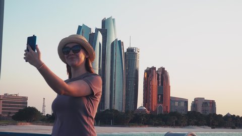 Young Caucasian Female Tourist Taking Selfie Pictures with Abu Dhabi Cityscape with Mobile Phone. Woman on Solo Travel Vacation in Arab Middle East Countries