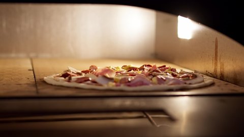 Italian chef putting raw cheese pizza into hot oven. Pizza maker man preparing tasty pizza with ham and mushrooms baking it in the oven. Food preparation. Appetizing footage. Close-up.