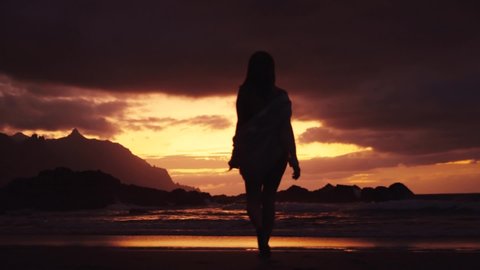 Back view of silhouette of a woman walking along the beach to the sea red sunset dramatic sky relax enjoying travel sexy waves cloudy slow motion