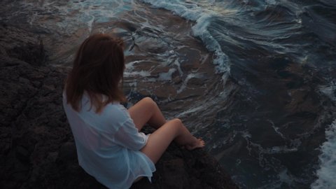 Close up top view of unrecognizable woman in swimsuit and white shirt sitting on rock in the sea and wathcing to big waves crashing agains the stone relaxing sunset beautiful travel alone slow motions