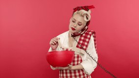 Excited pin up woman cooking and talking on telephone on pink background