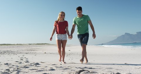 Front view of a happy Caucasian couple in love enjoying free time on the beach together, walking and holding hands with blue sky and sea in the background in slow motion