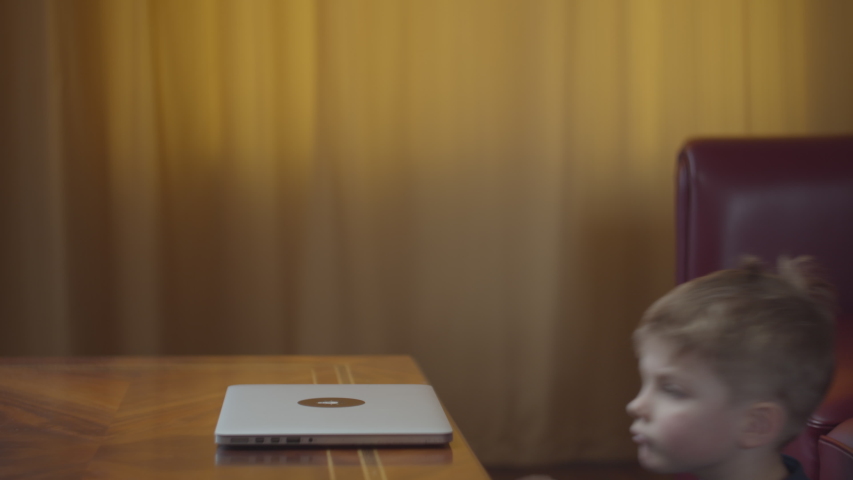 Blonde preschool boy opening and typing on laptop and smiling looking at computer screen. Kid learning online at home sitting at the table. Side view
