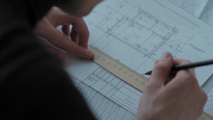 Architect works with blueprints: shot from behind a shoulder. Papers and drawings on the desk in front of the engineer. He draws the project using rulers and a pencil. Close up: hands of an architect Royalty-Free Stock Footage #1047256552