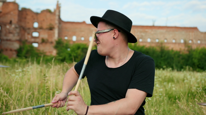 man musician drummer dressed in black clothes, glasses and hat, energetically playing on the drum set, on the street, near the destroyed building on a Sunny day, around tall green grass, slow motion