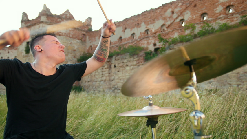 musician drummer dressed in black clothes with an earring in his ear, emotionally playing the drum set and cymbals, twists drumsticks, on the street near the destroyed building, slow motion