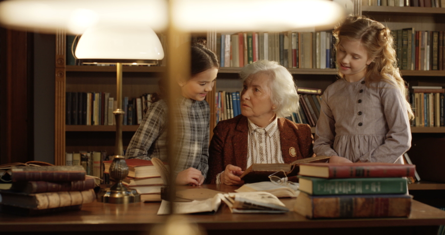 Caucasian old beautiful grandmother sitting at table in library and telling fairytale for two pretty cute small granddaughters. Little schoolgirls listening to female senior librarian reading book.