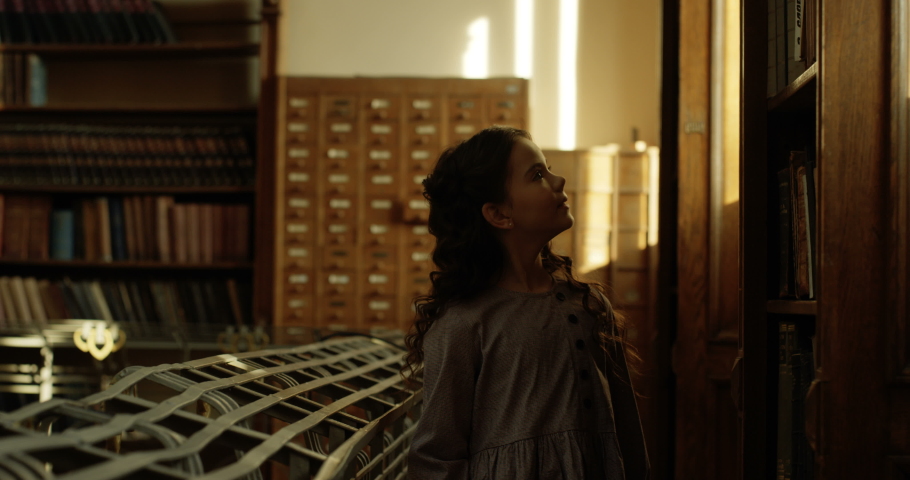 Caucasian beautiful small cute girl with long curly hair walking library dark passage with sun rays falling on her face. Curious little teen kid roaming magical atmosphere in bibliotheca.