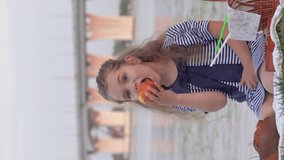 little cute child girl eating fresh fruit during picnic outdoors near river on background of bridge, smartphone vertical screen