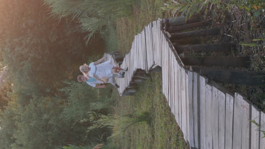 children recreation, cute kids friends boy and girl play catch-up and run on wooden bridge among green high vegetation in nature, smartphone vertical screen