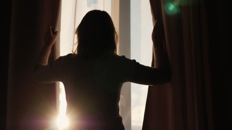 The silhouette of a woman who opens the curtains on the window and looks forward
