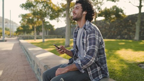Portrait of a smiling young man with earphone in his ears texting messages on mobile phone after riding on skateboard in the park