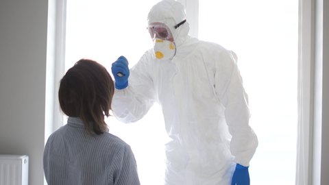 Young epidemiologist doctor in a white protective insulating suit examines a woman sitting on the bed. Coronavirus vaccine Covid19