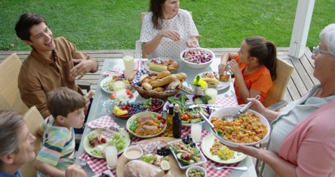 High angle view of a multi-generation Caucasian family outside at a dinner table set for a celebration meal, sitting, talking, standing and serving food, slow motion