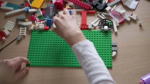 schoolgirl with tender hands constructs lego elements on green platform lying among colorful blocks close upper view