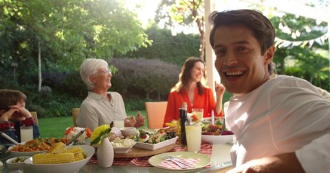 Side view of a Caucasian man sitting outside with his multi-generation family at a dinner table set for a meal, turning and smiling to camera, slow motion