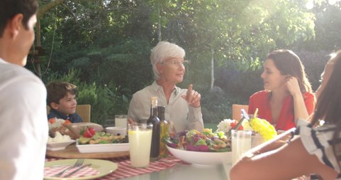 Front view of a senior Caucasian woman, her adult daughter and husband, grandson and granddaughter sitting outside at a dinner table set for a family meal, talking and smiling, backlit, slow motion