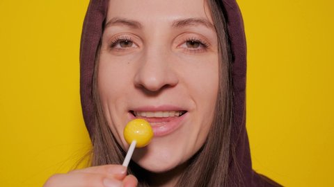 Beautiful young woman in a red hood sucks a yellow lollipop with a white wand. Female sexy lips suck a big yellow round lollipop. Copy space yellow background
