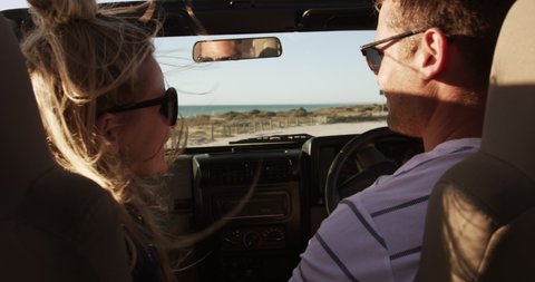 Rear view of a Caucasian couple enjoying free time during road trip together, sitting inside an open top car, driving and smiling with blue sky and sea in the background in slow motion 