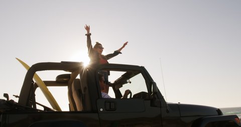 Side view of a happy Caucasian woman enjoying free time during road trip, standing in an open top car, with arms in the air wearing sunglasses smiling with blue sky and sea in the background in slow
