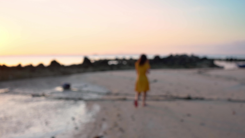 A girl in a yellow dress walks along the sandy shore of the ocean at sunset.