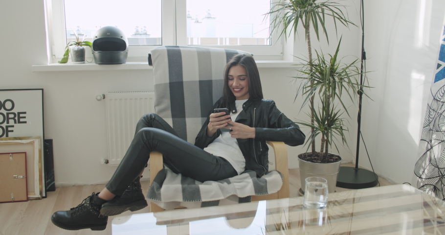 Young motorcyclist woman in black leather jacket woman sitting in a chair at home. Urban biker girl resting and using mobile phone . 4K video shooting by handheld gimbal