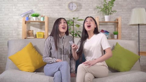 two happy asian girls catch flying dolar banknotes while sitting at home on the sofa in the living room slow mo