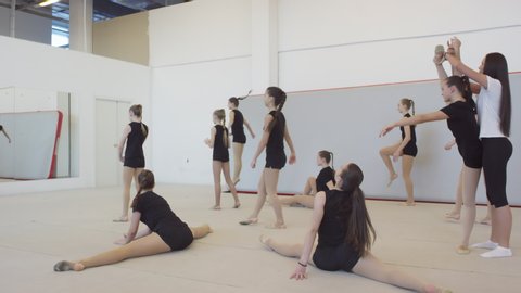 Tracking shot of teen and preteen girls in black t-shirts and shorts stretching and warming up during cheerleading or gymnastics practice. Young female coach helping one of students