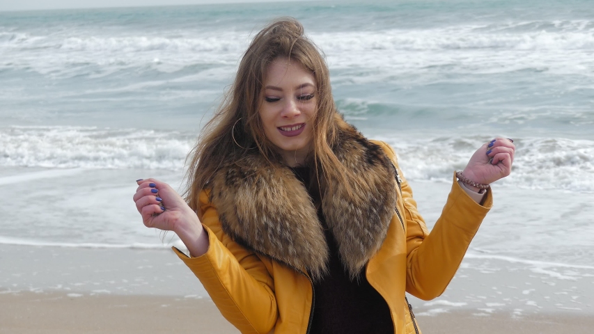 4K video Young beautiful European-style girl on the seashore in a burgundy skirt and yellow jacket slowly spills sand out of her hands, swirls and dances