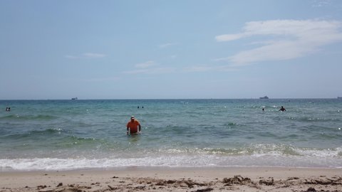 Ft. Lauderdale, Florida - May, 2019: An old man wearing a bathing suit and an orange shirt is seen trying to keep his balance as he exits the ocean onto the beach on a warm sunny day 